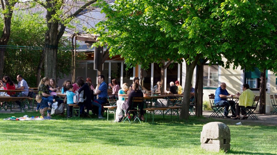 Gastgarten mit Biertischen unter alten Bäumen, im Hintergrund das Wirtschaftsgebäude